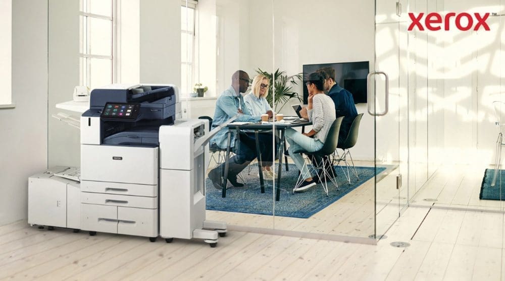 Xerox® Smart Start : intégration transparente au bureau Une imprimante multifonction Xerox AltaLink B8245 moderne trône au premier plan d'un bureau lumineux. À l'arrière-plan, un groupe de personnes discutent autour d'une table.