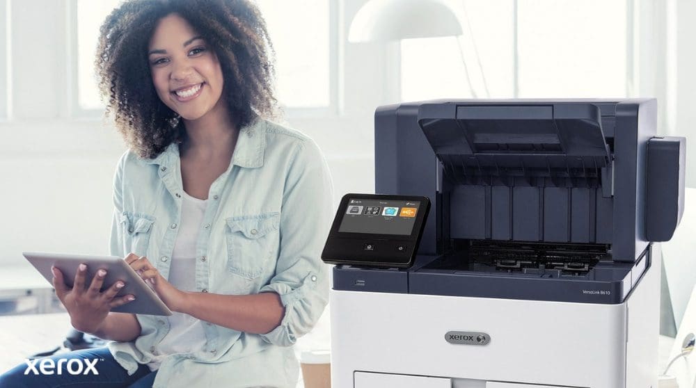 Xerox® Workplace Cloud et Suite : intégration conviviale Une personne souriante est assise à côté d'une imprimante Xerox VersaLink B610 blanche, tenant une tablette et regardant la caméra, soulignant la nature conviviale des solutions Xerox.