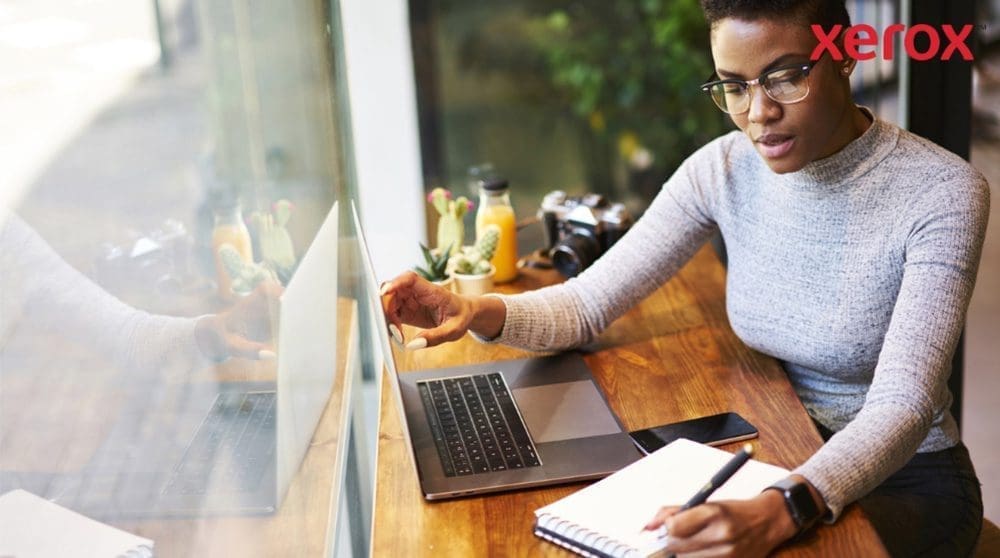 Xerox® Workplace Cloud et Suite : productivité améliorée Une personne portant des lunettes travaille à une table en bois, regarde un écran d'ordinateur portable et écrit dans un cahier, avec un smartphone à proximité, suggérant un environnement de travail productif
